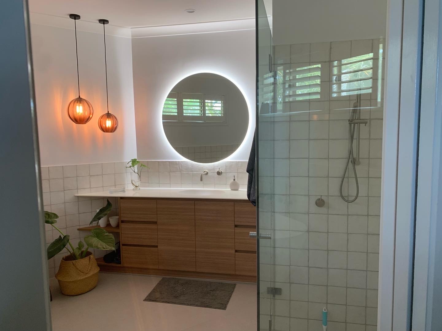 A finished bathroom with a timber vanity, round backlit mirror, pendant lights and a glass shower