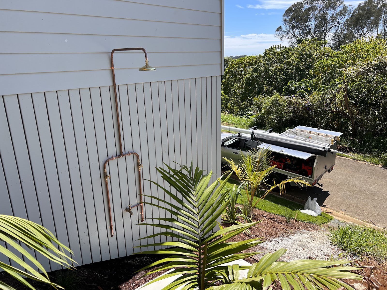 An outdoor copper shower mounted on a white weatherboard wall, surrounded by palms